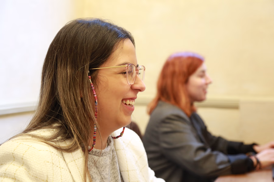 Two women sitting and smiling, one with glasses and long hair in the foreground, and one with red hair in the background.