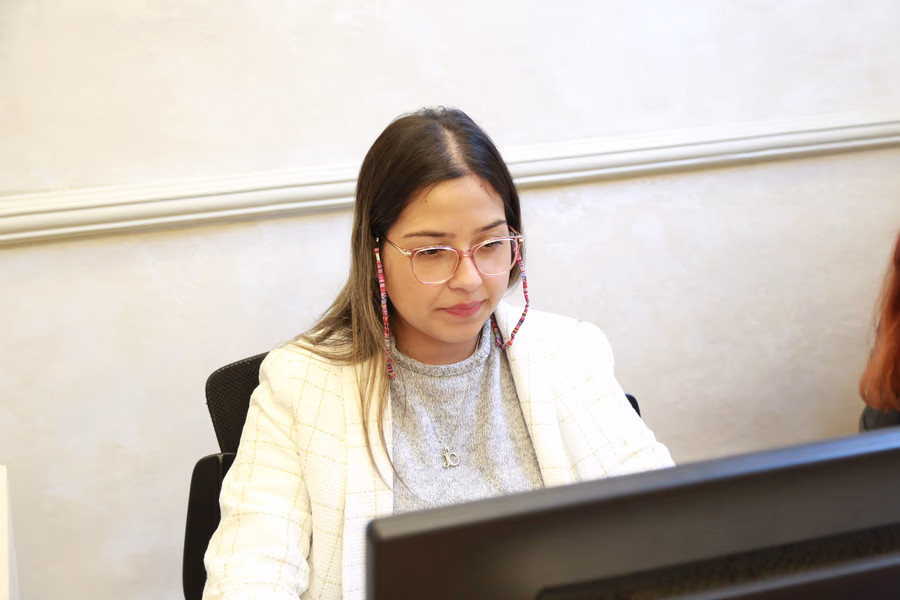Woman wearing glasses and a light-colored blazer working on a desktop computer in an office setting.