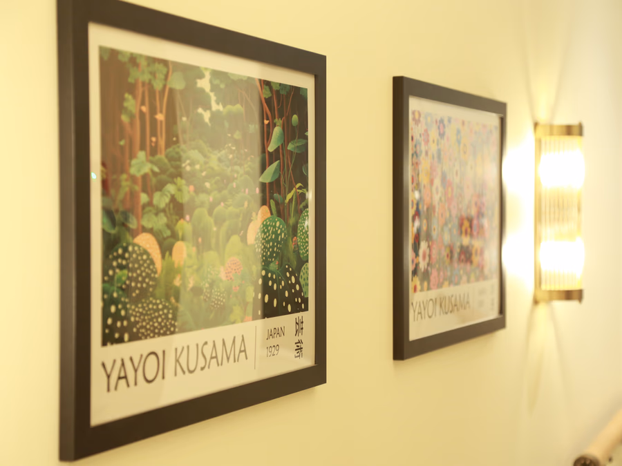 Two framed posters by Yayoi Kusama with forest and floral themes hanging on a beige wall next to a lit wall sconce.