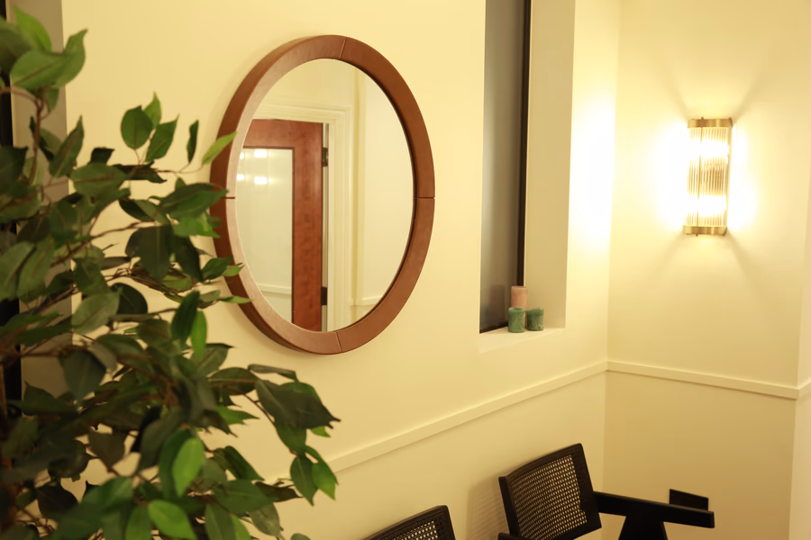 Indoor waiting area with a round wooden-framed mirror, a green plant, two black chairs, and a wall sconce light.