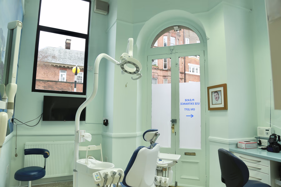 Dental clinic room with dental chair, equipment, window, and glass door with 'Please use entrance on left' sign.