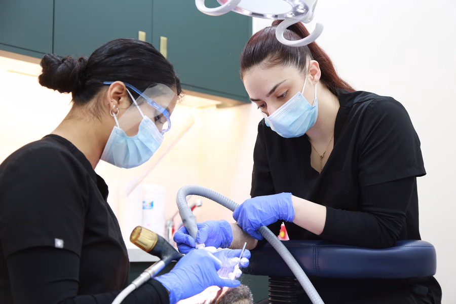Two female dental professionals in black scrubs, masks, and gloves performing a dental procedure on a patient.