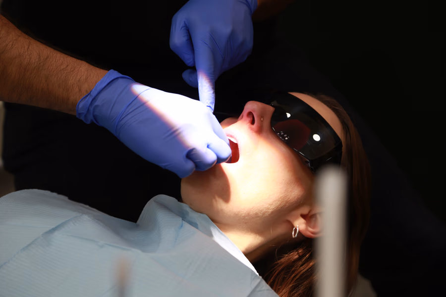 Dental care professional wearing blue gloves examining a patient's mouth under a bright light.