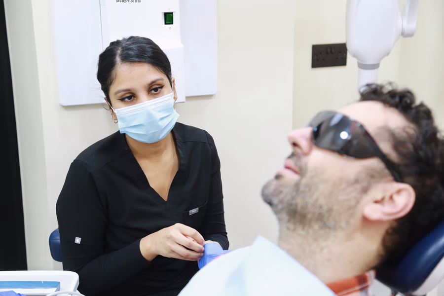 Dentist wearing a mask and gloves preparing to treat a male patient reclining in a dental chair, wearing protective glasses.