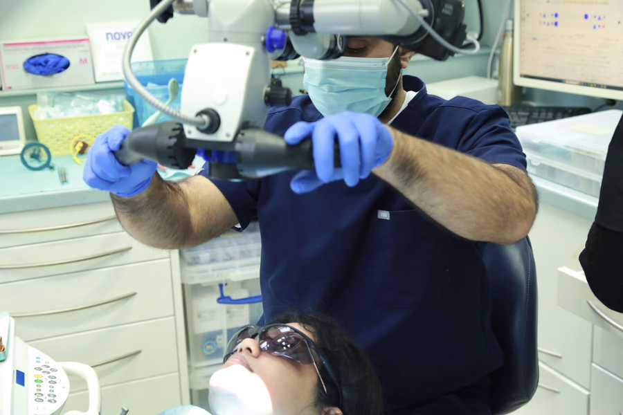 Dentist wearing blue gloves and a mask using a dental microscope while examining a patient wearing protective glasses.