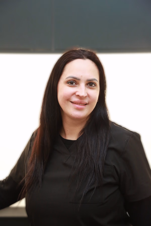 Woman with long dark hair wearing a black shirt, smiling at the camera against a blurred light background.