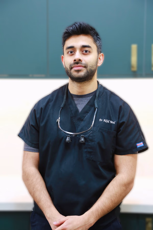 Male dentist wearing black scrubs and dental loupes around his neck, standing in a modern clinic.
