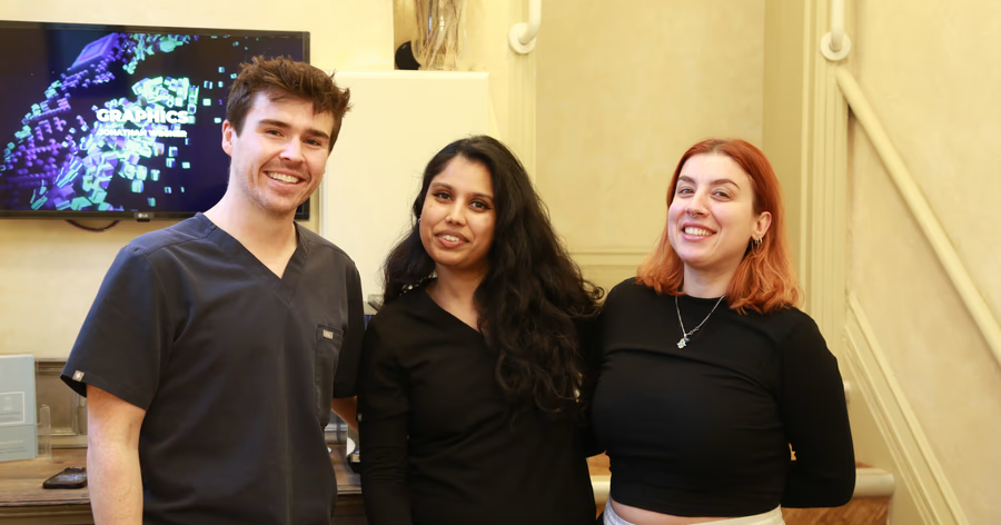 Three smiling young adults standing indoors in front of a counter and a screen displaying the word 'GRAPHICS'.