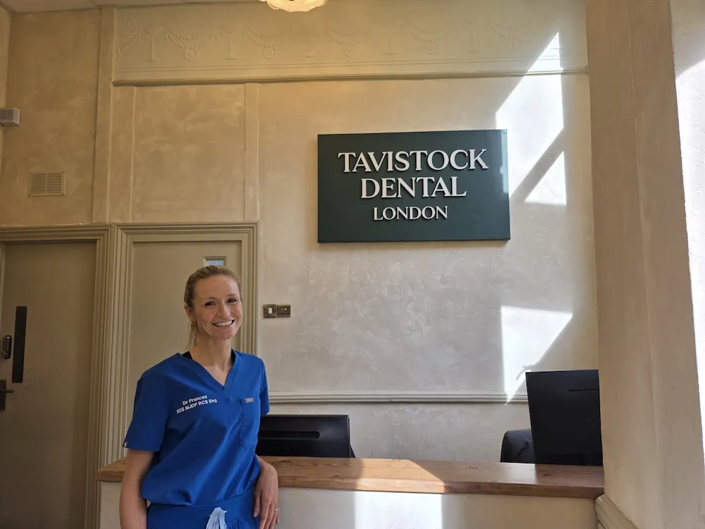 Smiling female dentist in blue scrubs standing beside the reception desk at Tavistock Dental London clinic.