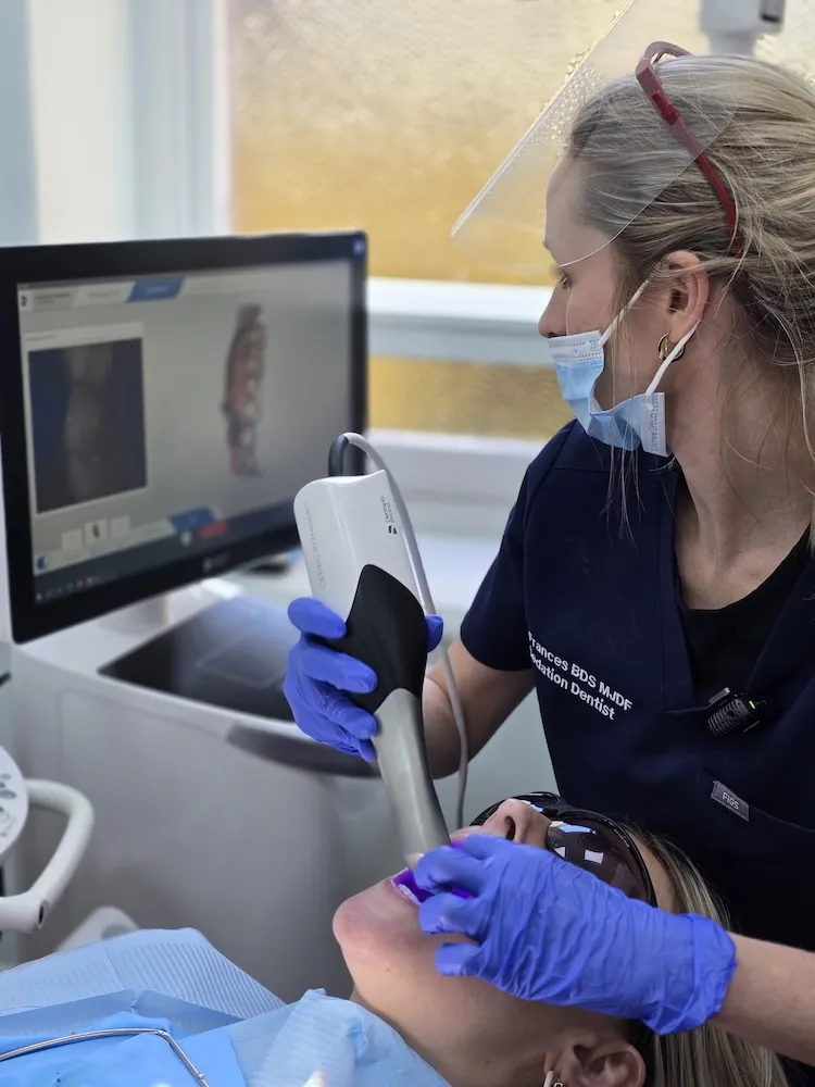 Dentist wearing a face mask and face shield uses an intraoral scanner on a patient reclining in a dental chair, with a computer screen showing a 3D dental image in the background.