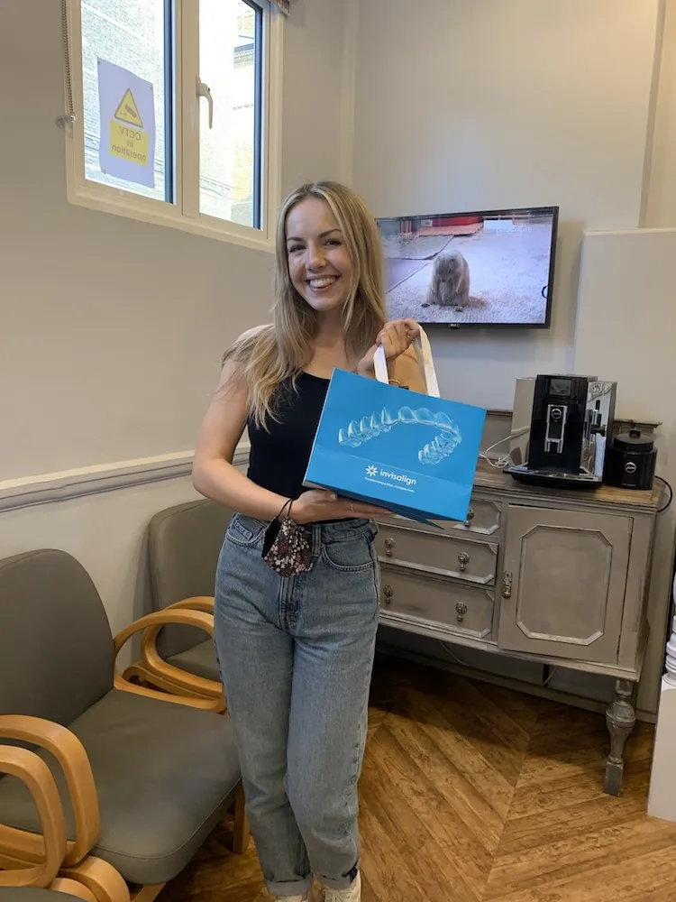 Smiling woman holding a blue Invisalign bag in a waiting room with chairs and a cabinet.