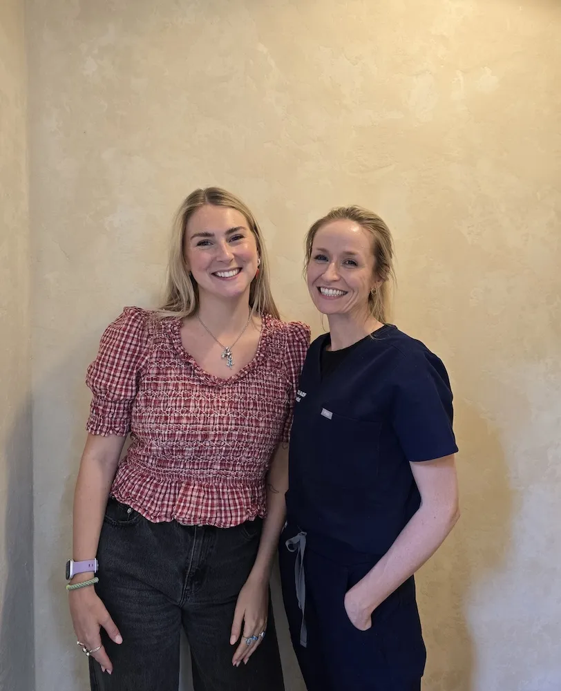 Two smiling women standing side by side against a beige textured wall, one wearing a red checkered blouse and the other in navy blue scrubs.