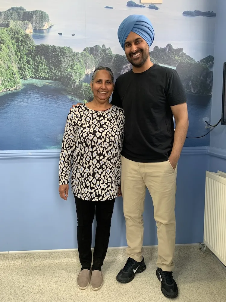 Smiling man wearing a blue turban and black shirt standing next to a smiling woman in a floral blouse against a wall mural of a tropical island landscape.