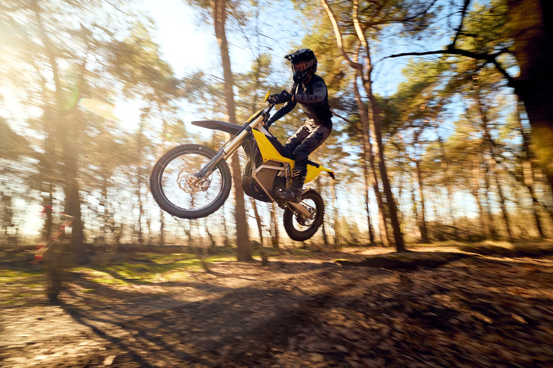 Person wearing black gear and helmet jumping with a yellow dirt bike in a sunlit forest.