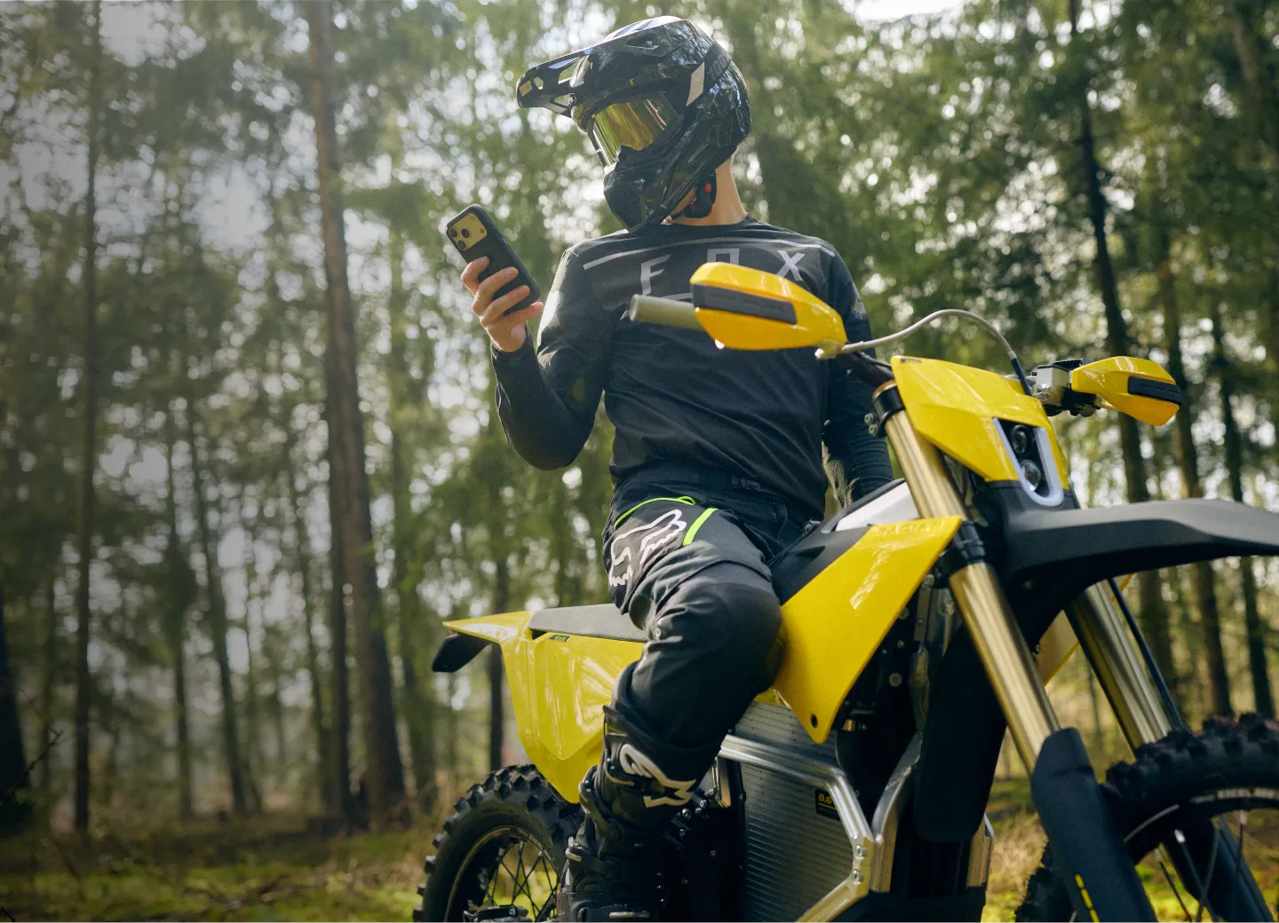 Person wearing black motocross gear and helmet sitting on a yellow dirt bike looking at a smartphone in a forest setting.