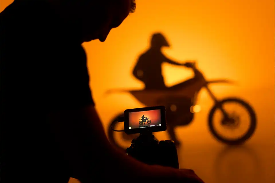 Silhouette of a person filming a motorcyclist on an orange background with the motorcycle visible on the camera screen.