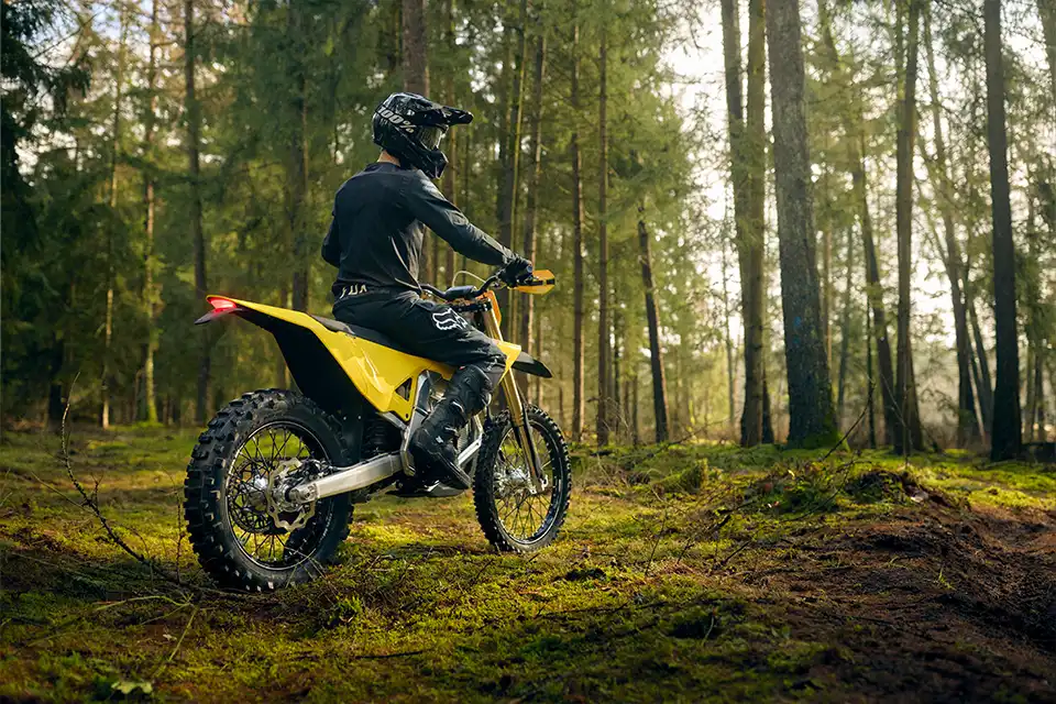 Person wearing black gear and helmet riding a yellow dirt bike in a sunlit forest.