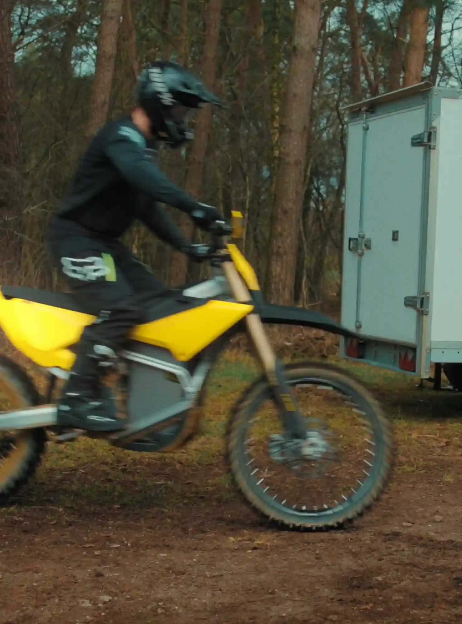 Person wearing black helmet and gear riding a yellow dirt bike on a trail with trees and a white trailer in the background.