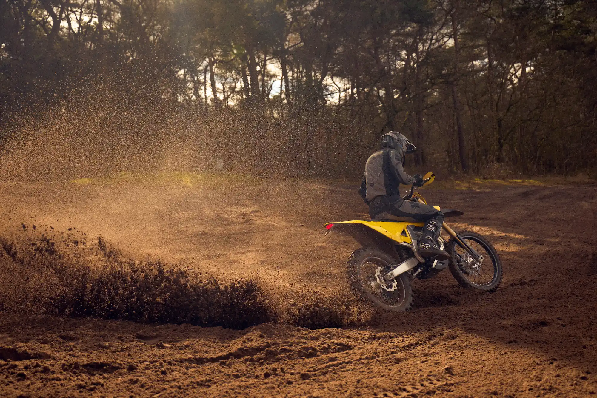 Motorcyclist in protective gear riding a yellow dirt bike, kicking up mud on a forest trail.