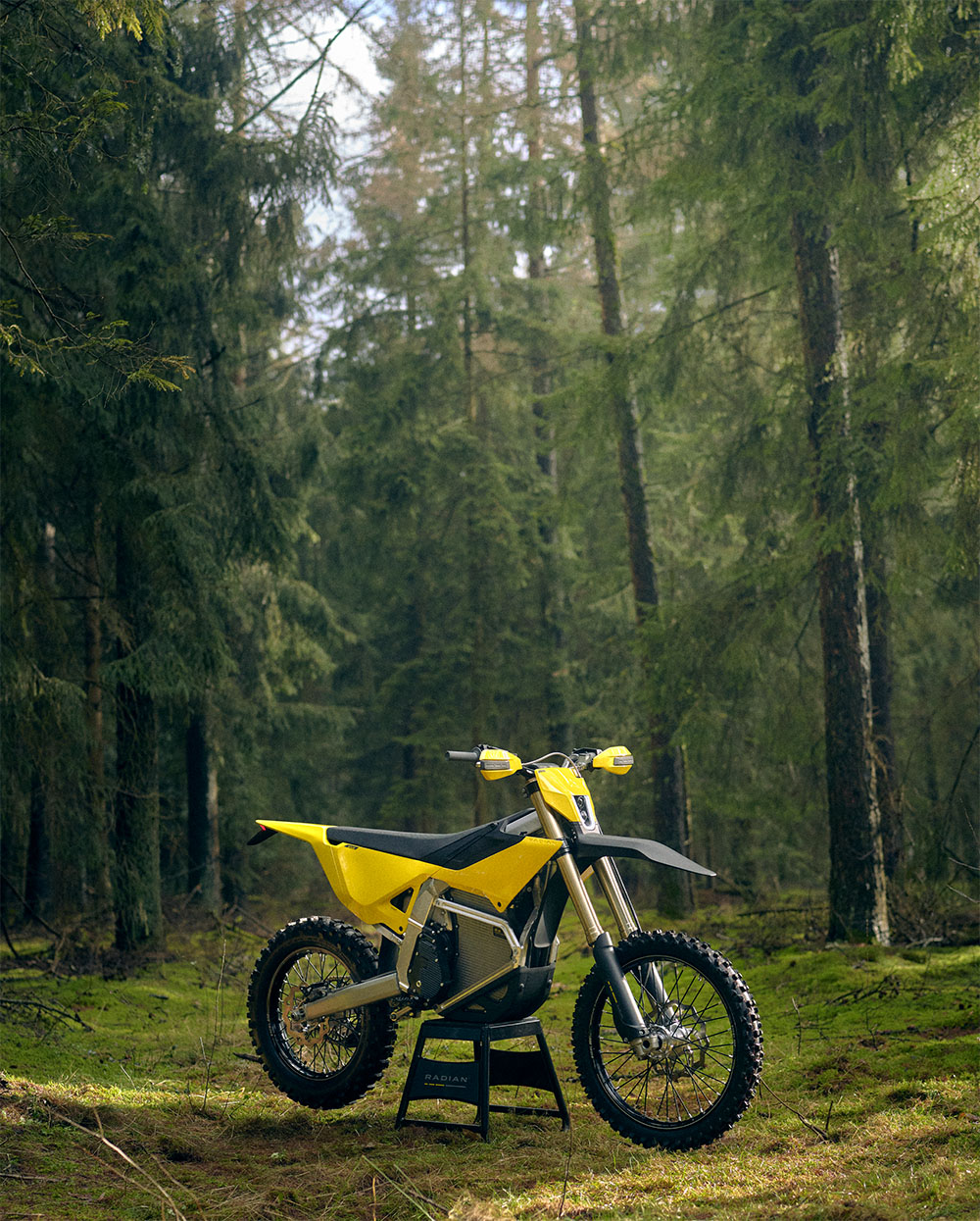 Yellow and black off-road motorcycle standing on a stand in a forest with green mossy ground.