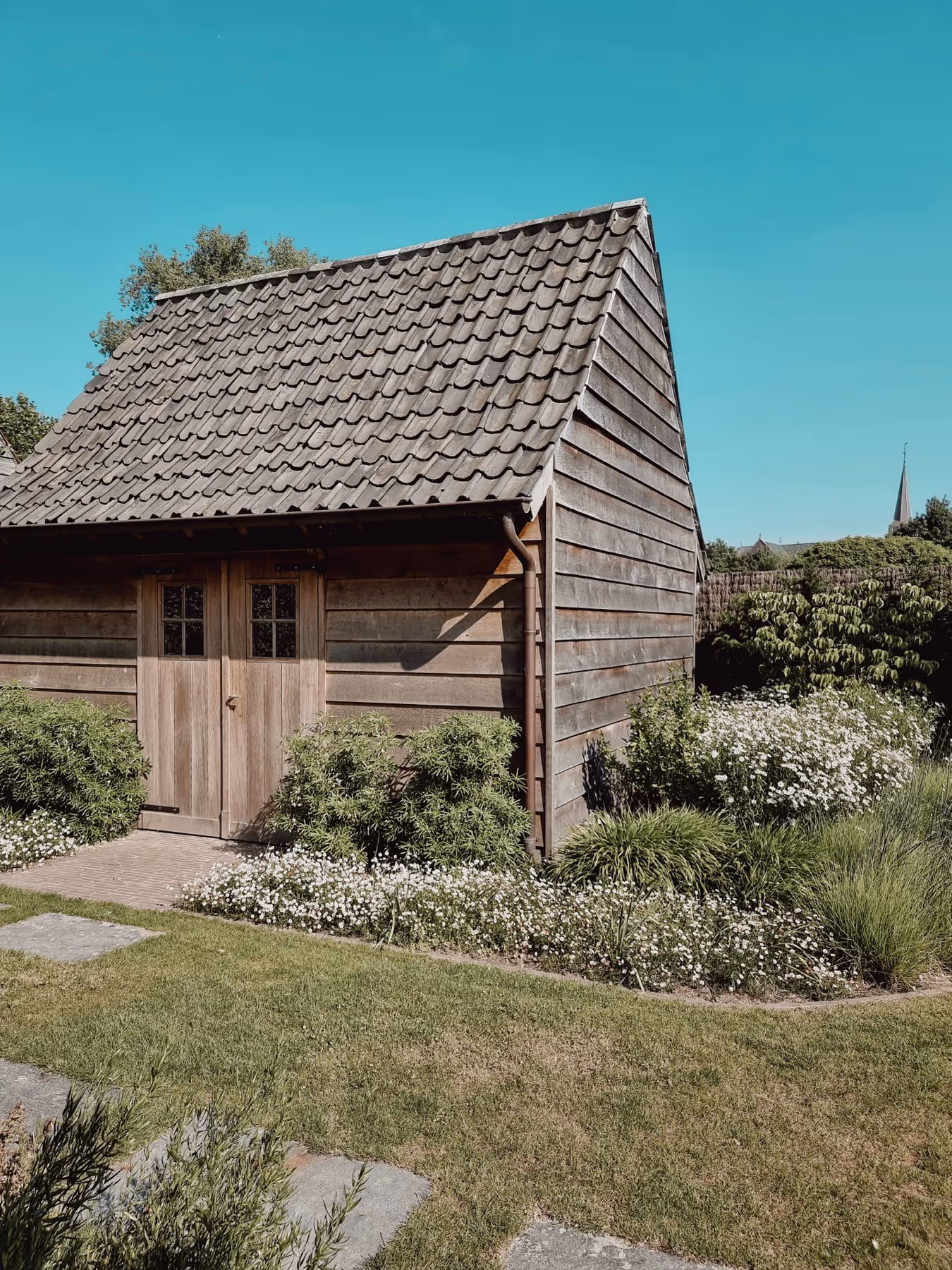 Wooden garden shed with tiled roof surrounded by green bushes, white flowers, and a grassy lawn under a clear blue sky.