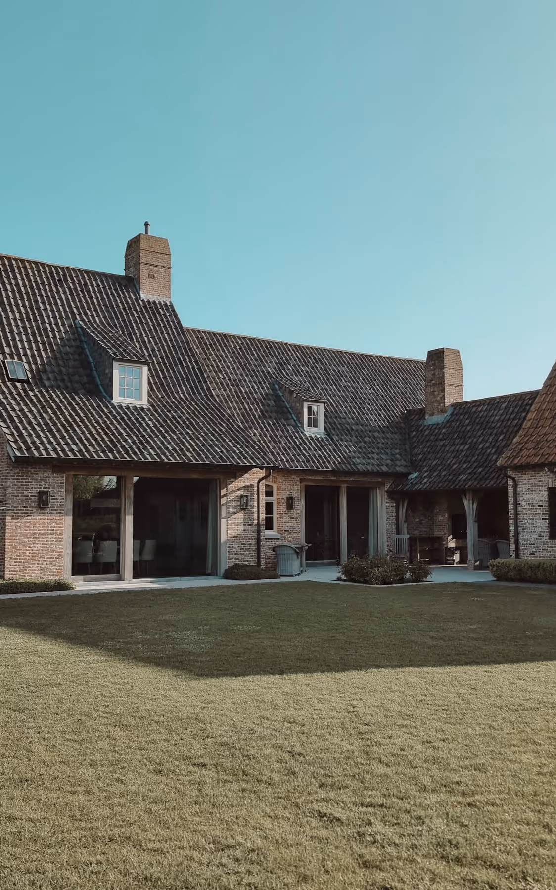 Traditional brick house with dark tiled roof, multiple chimneys, large sliding glass doors opening to a patio and a spacious green lawn.