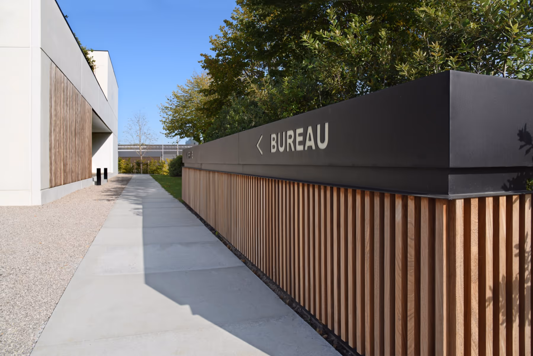Outdoor walkway beside modern building with wooden paneling and a sign pointing left labeled 'BUREAU'.