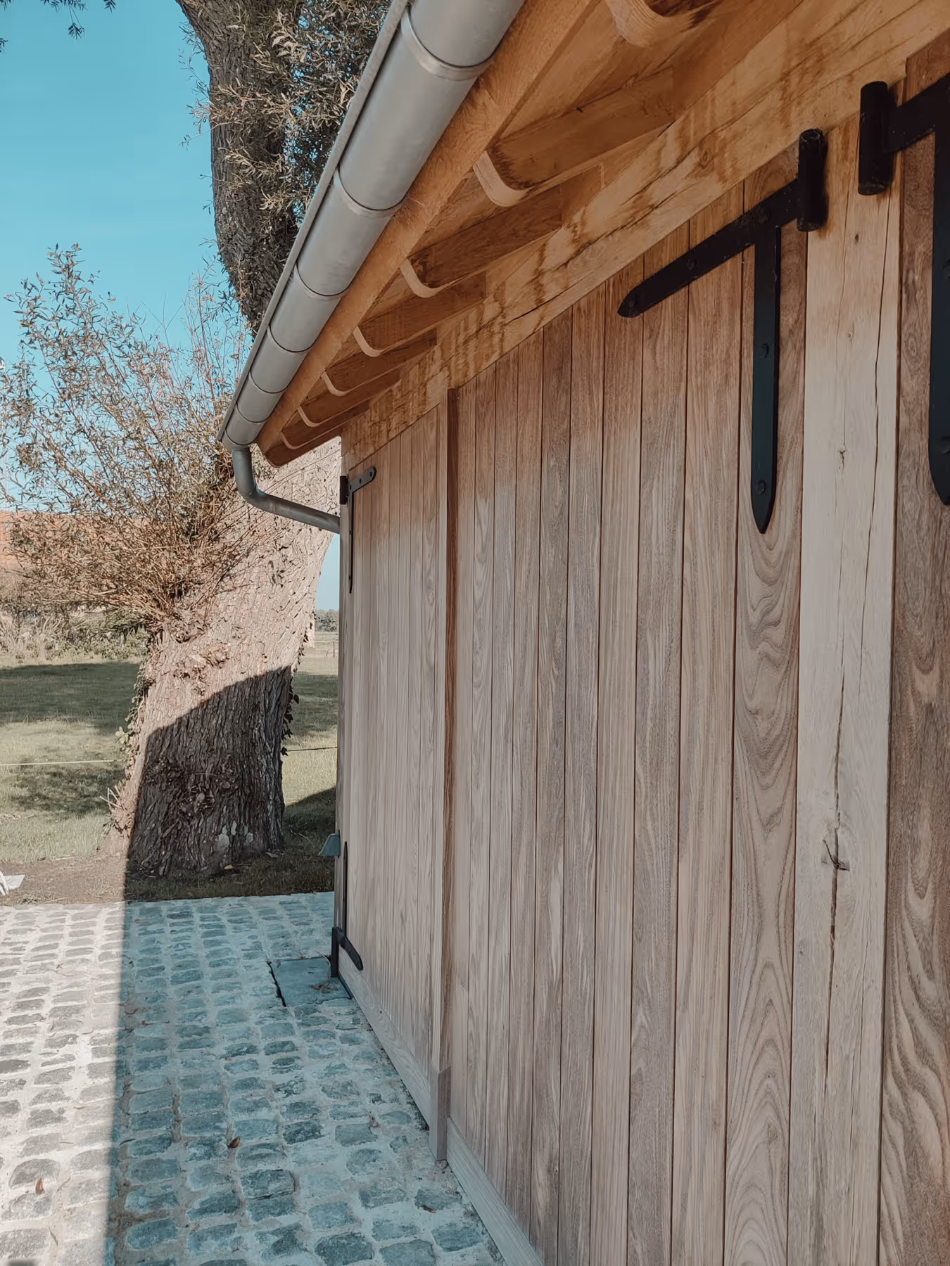 Wooden barn door with black metal hinges next to a large tree on a cobblestone path under a wooden roof.