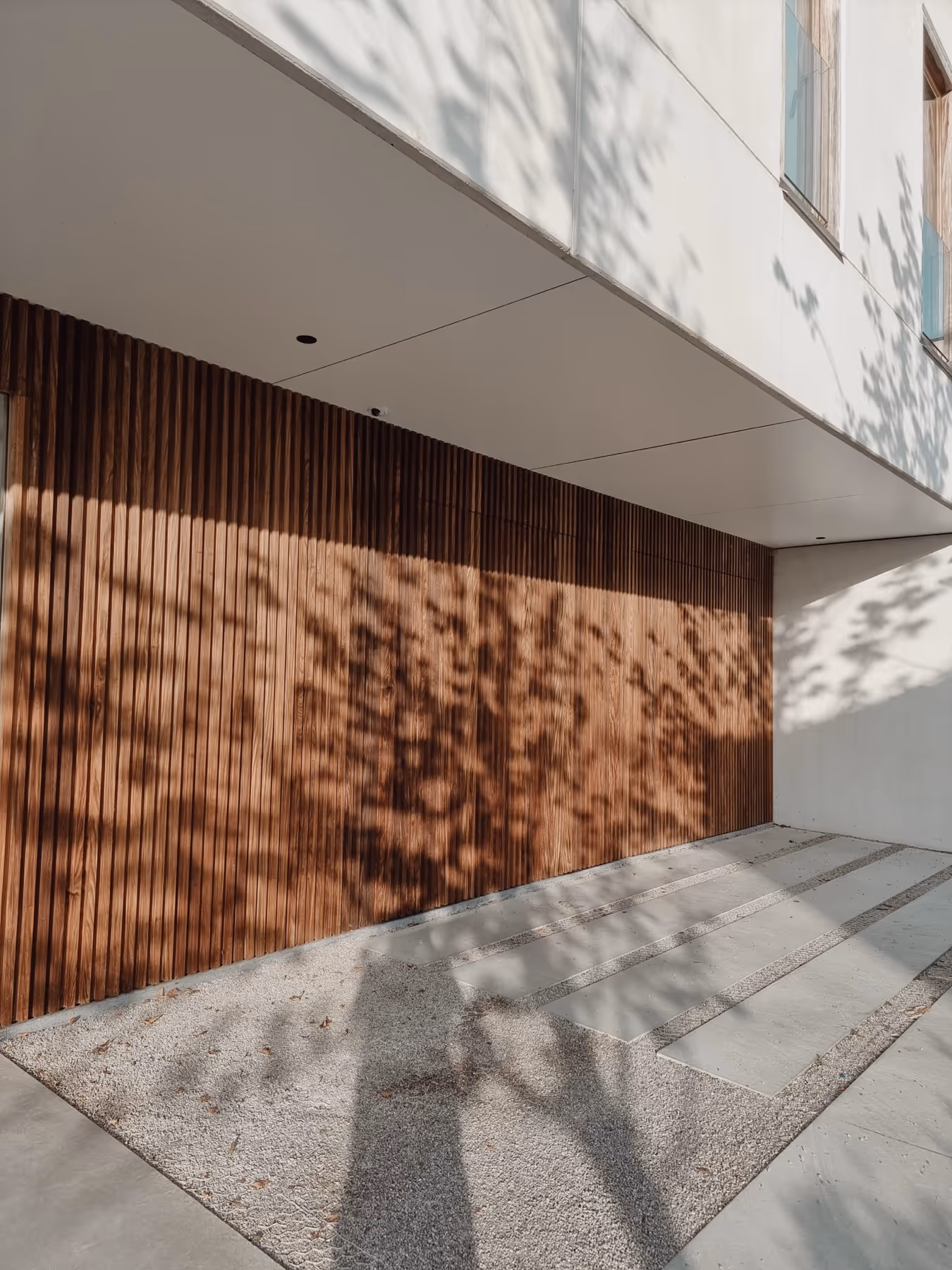 Modern exterior with vertical wooden slats wall, shadows from nearby trees, and a concrete path with gravel in between.