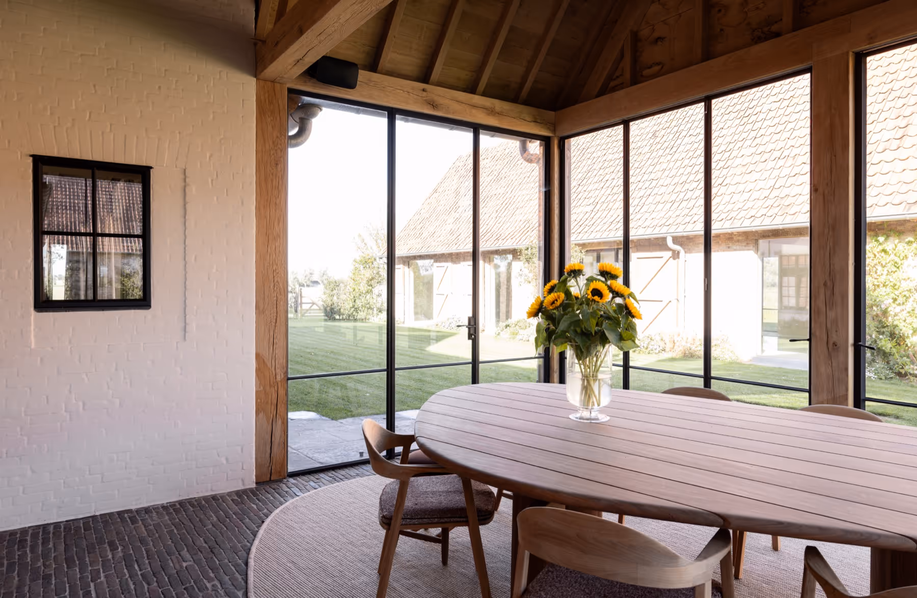 Covered terrace with a wooden oval table, six chairs, and a vase of sunflowers, overlooking a garden through large glass doors.