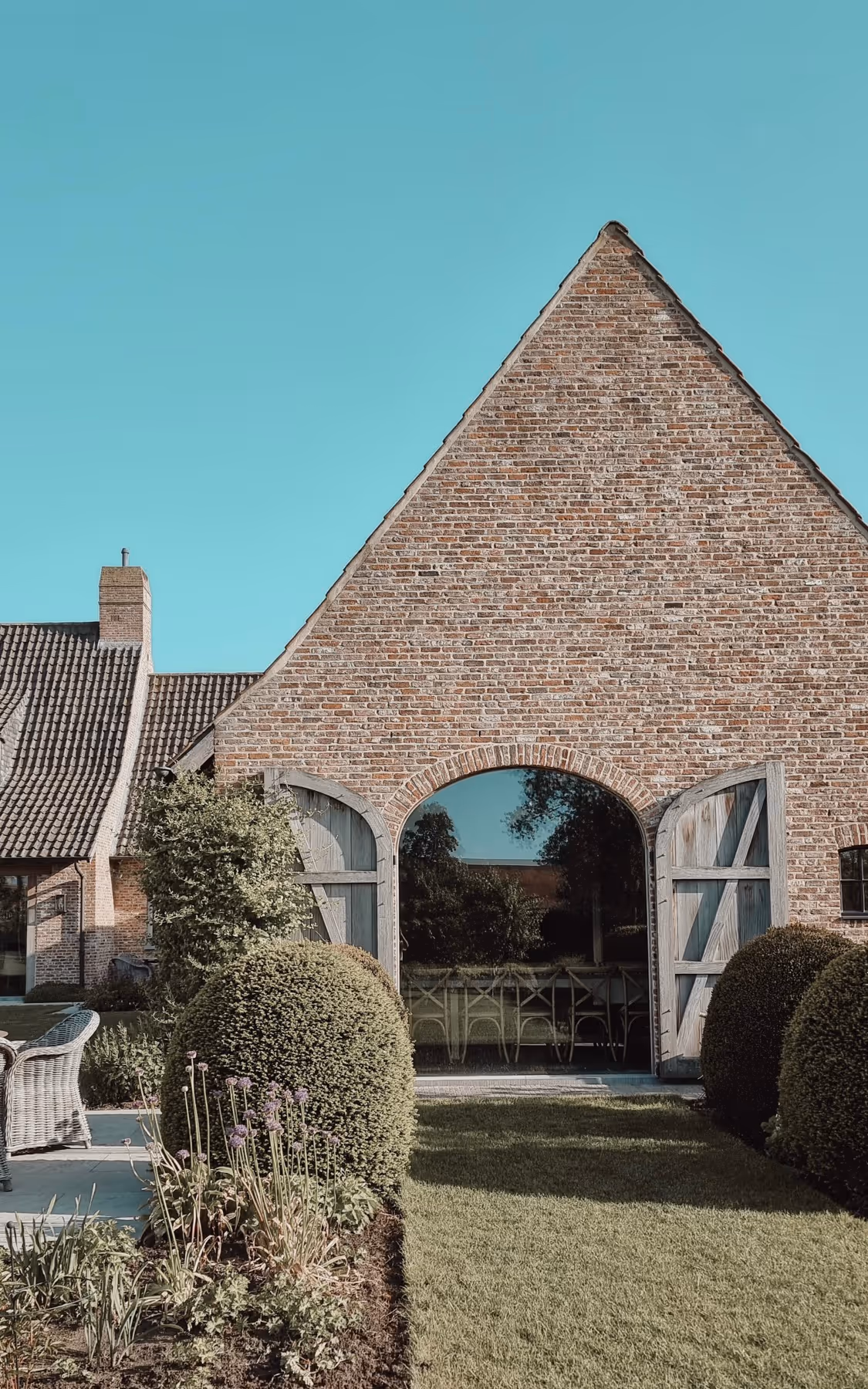 Brick house with a large arched glass door flanked by open wooden shutters and surrounded by manicured bushes and garden.