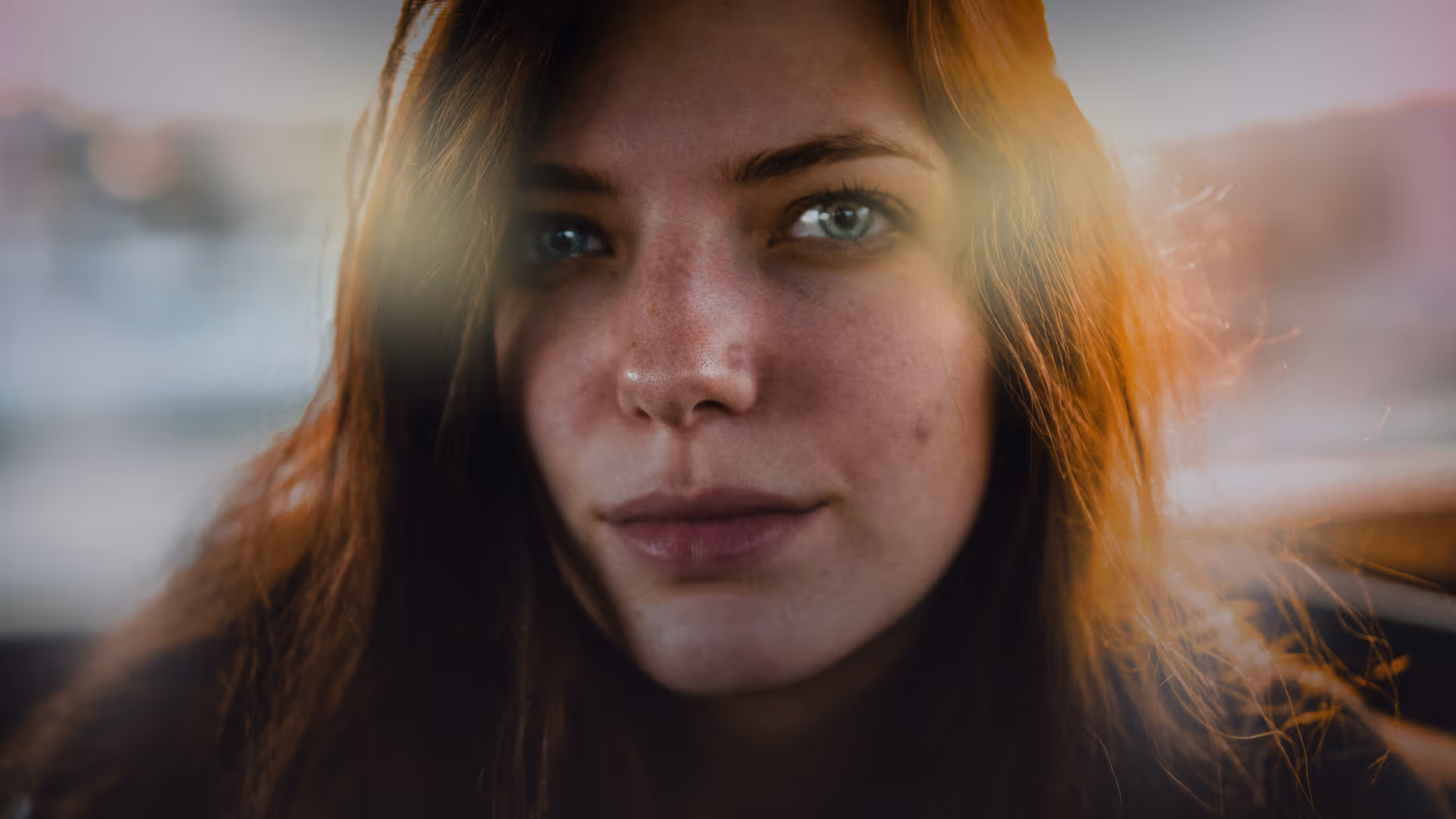 Close-up of a young woman with long brown hair and blue eyes, softly lit by warm sunlight.