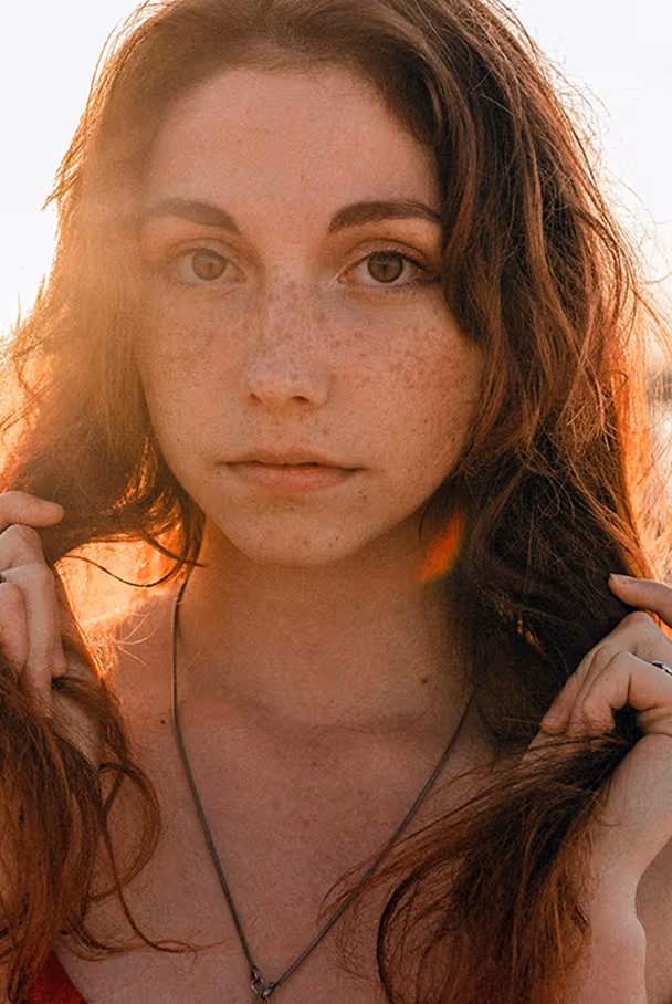 Close-up of a young woman with long brown hair and freckles, holding her hair with both hands, backlit by warm sunlight.