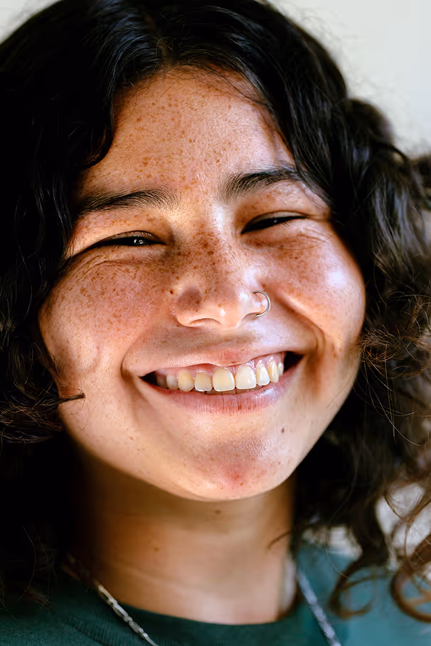 Close-up of a young person with curly dark hair, freckles, nose ring, and a broad smile showing teeth.