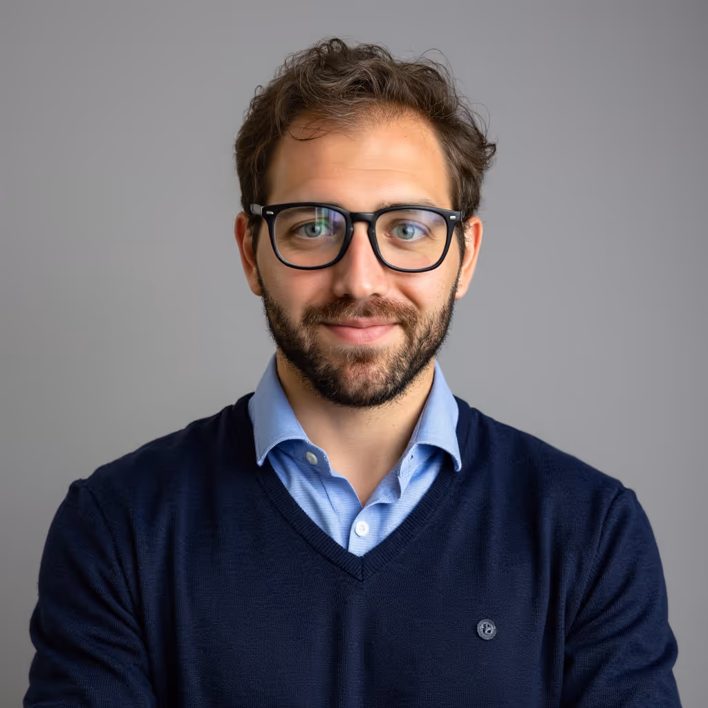 Smiling man with glasses, beard, wearing a blue sweater over a light blue collared shirt, standing with arms crossed.