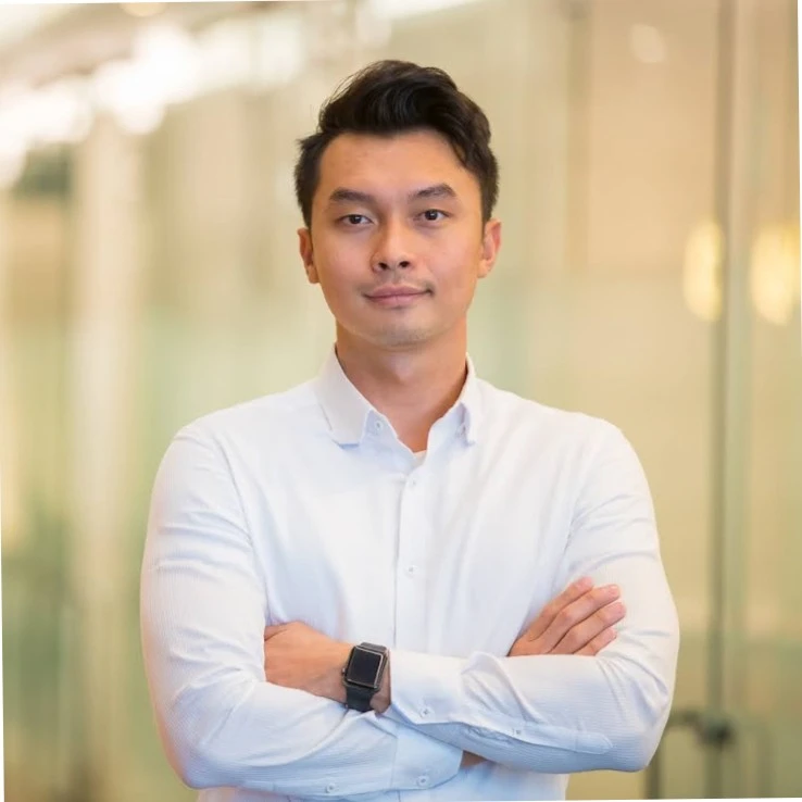 Man with short dark hair wearing a white button-up shirt and a smartwatch, standing with arms crossed in an office setting.