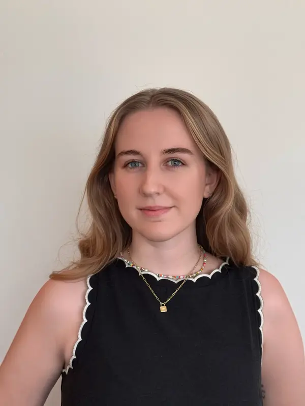 Young woman with long blonde hair, blue eyes, and wearing a black sleeveless top with white scalloped edges, smiling slightly against a plain light background.