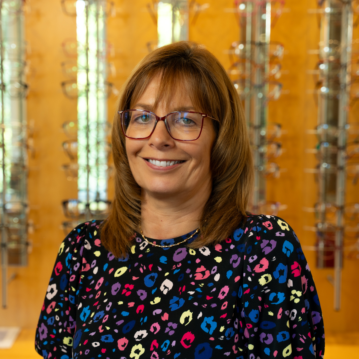 Smiling woman wearing glasses and a colorful patterned top with eyeglasses display racks behind her.