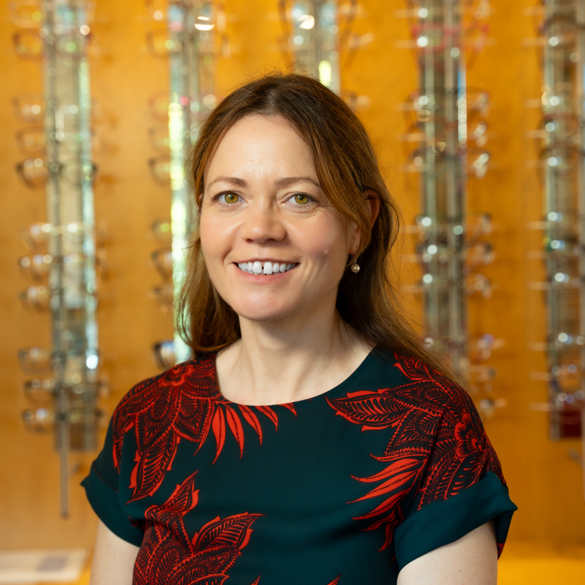 Smiling woman with brown hair in a red and green patterned shirt stands in front of eyeglass displays.