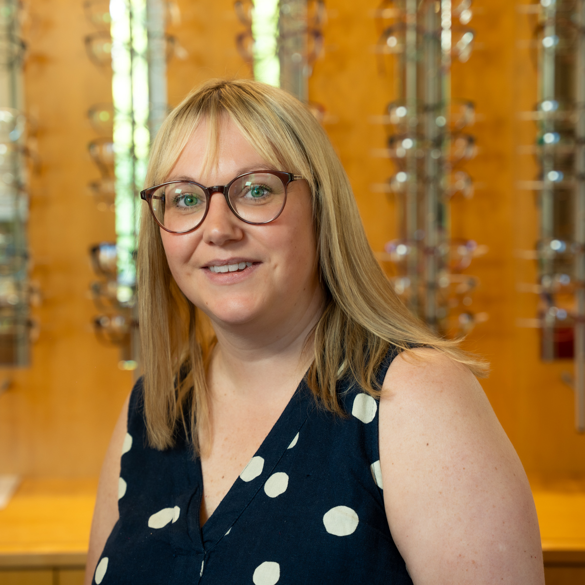 Smiling woman with blonde hair wearing glasses and a navy blue dress with white polka dots.