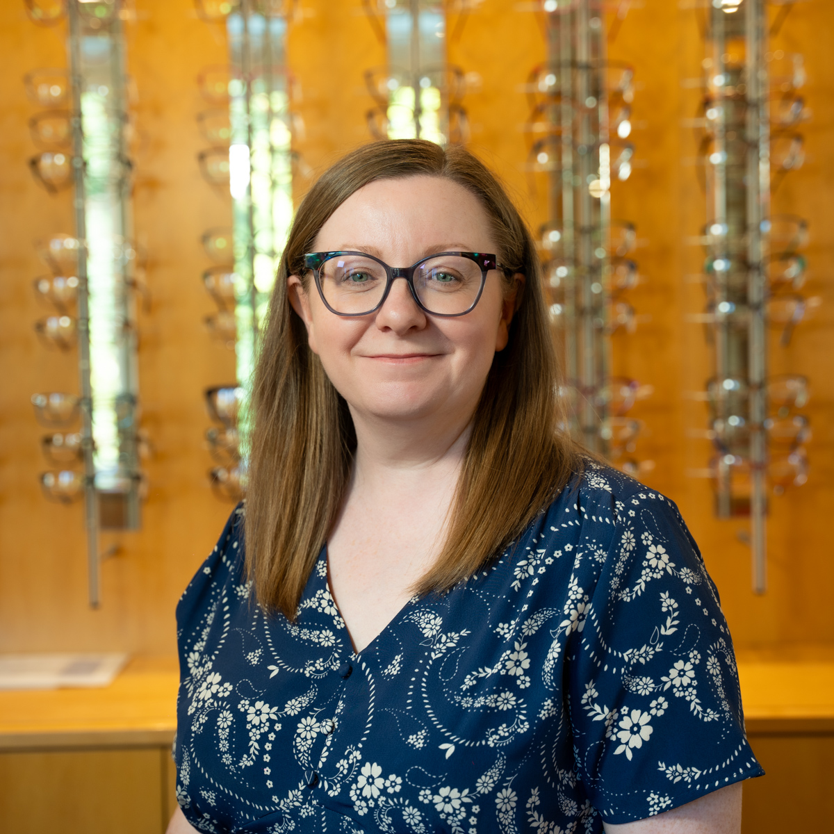 Smiling woman with straight brown hair and glasses in front of eyeglasses display wall.