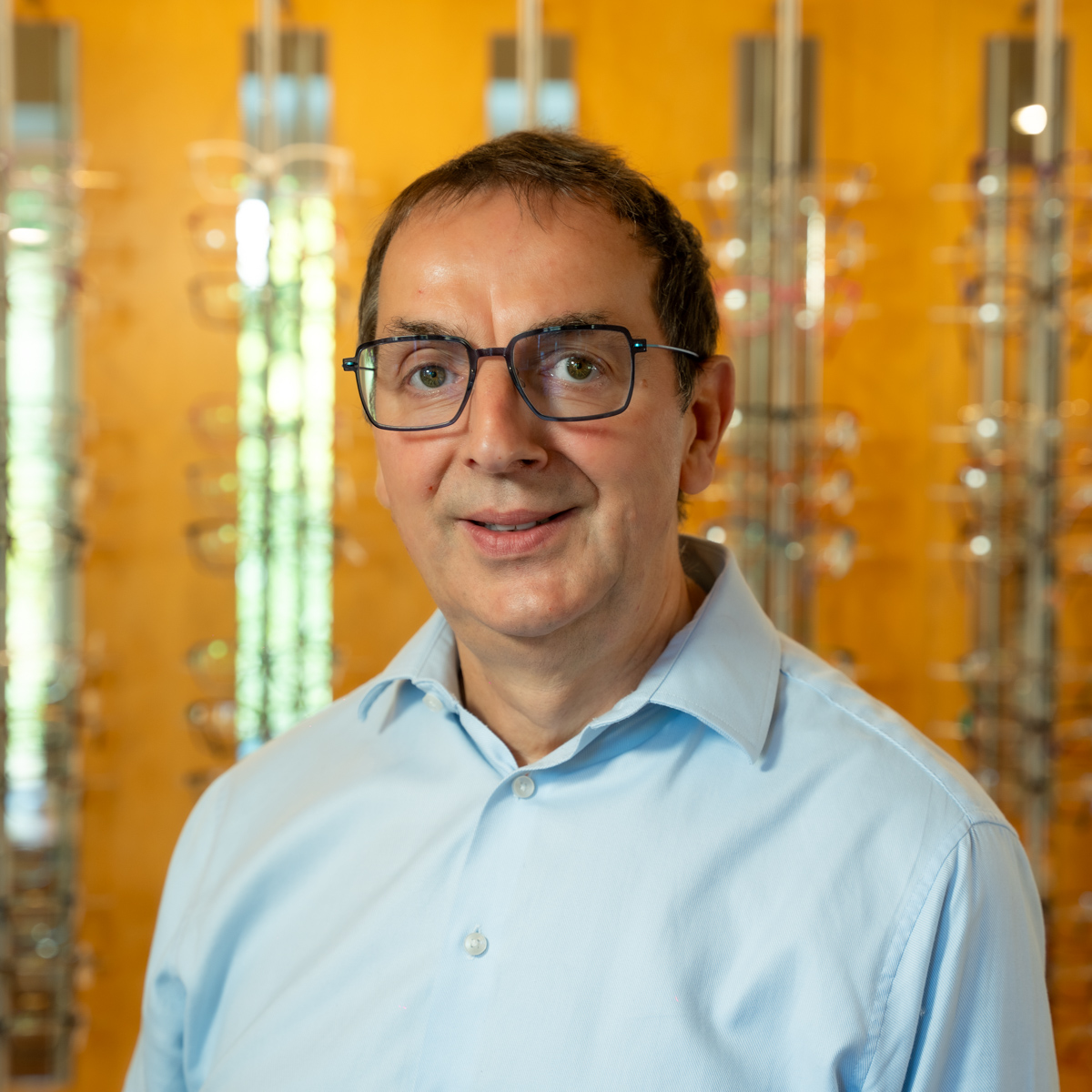 Middle-aged man wearing glasses and a light blue shirt standing in front of an eyewear display.