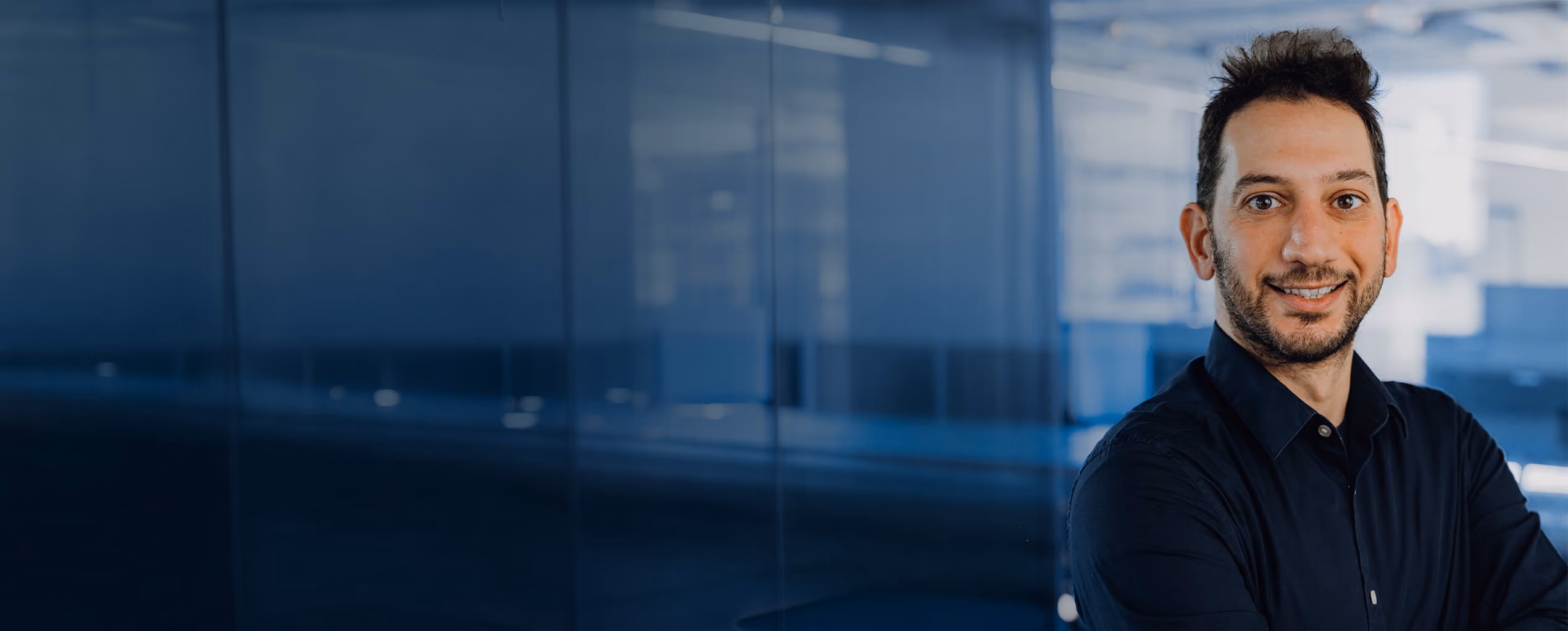 Smiling man in a dark shirt standing with arms crossed in a modern office.