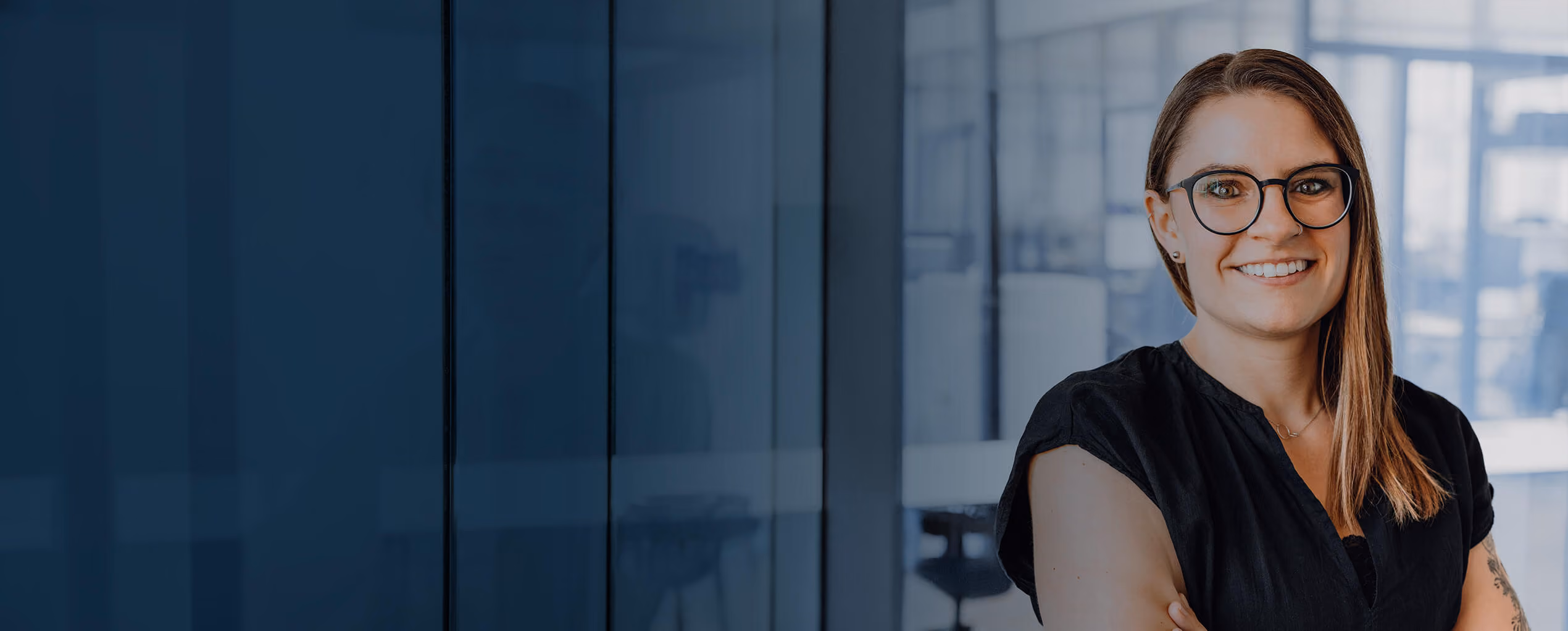 Smiling woman with glasses stands with arms crossed in a modern office.