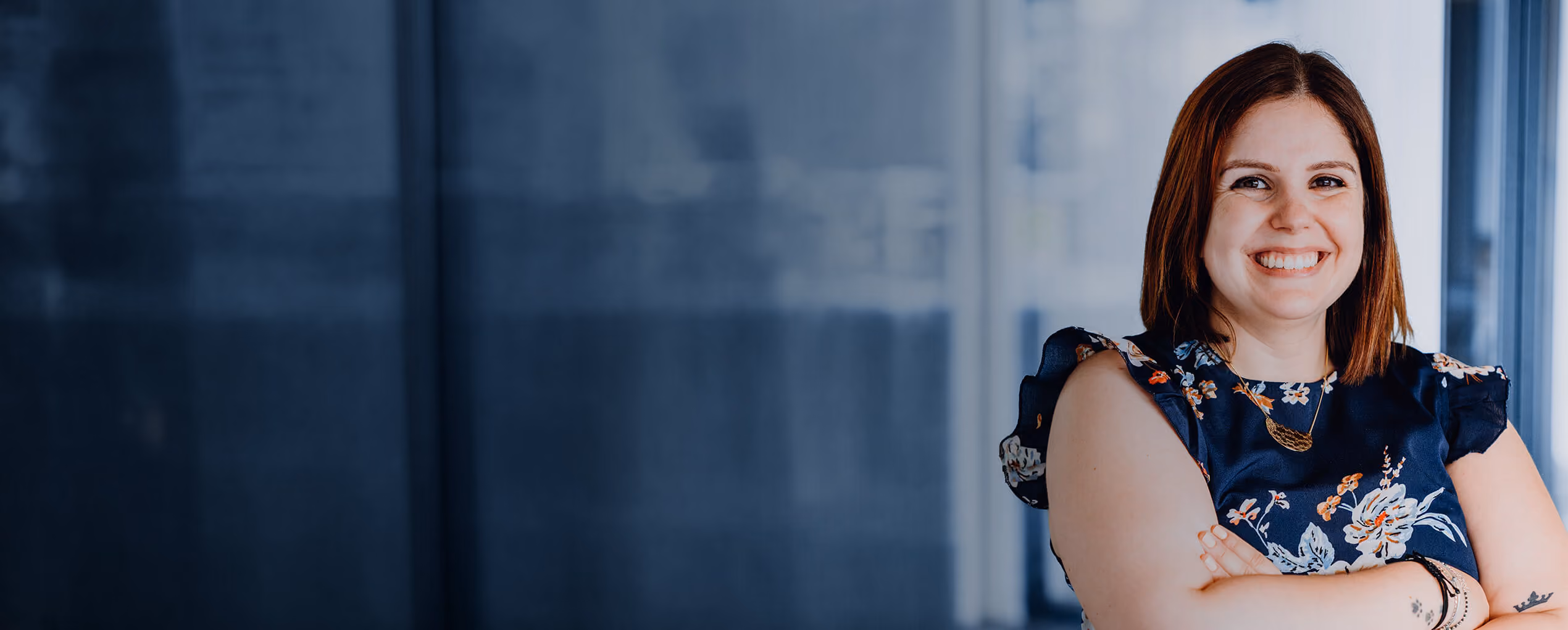 Smiling woman with brown hair wearing a blue floral dress standing with arms crossed.