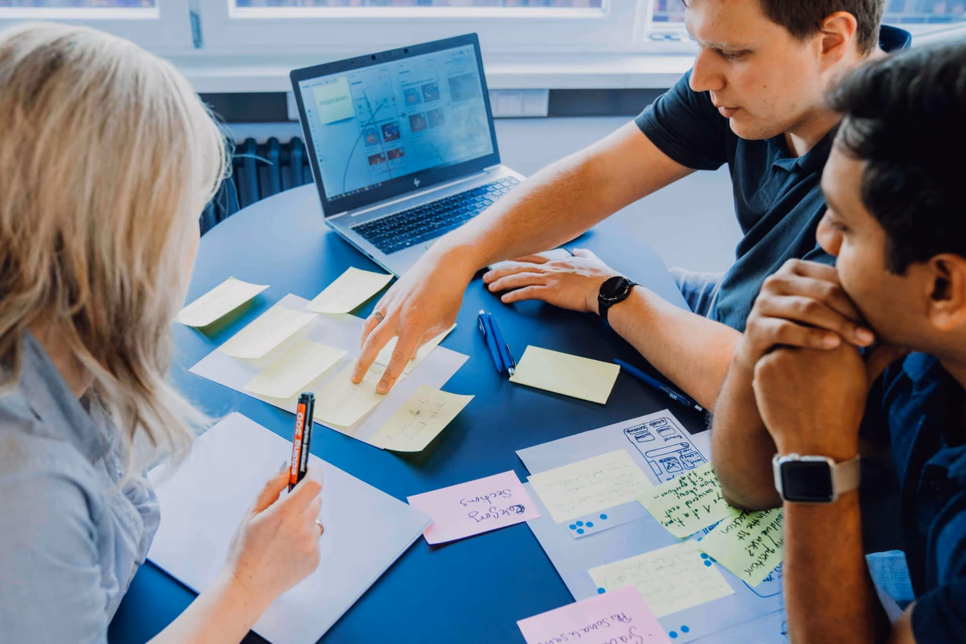 Three people discussing notes and plans on sticky notes and papers at a round table with a laptop.