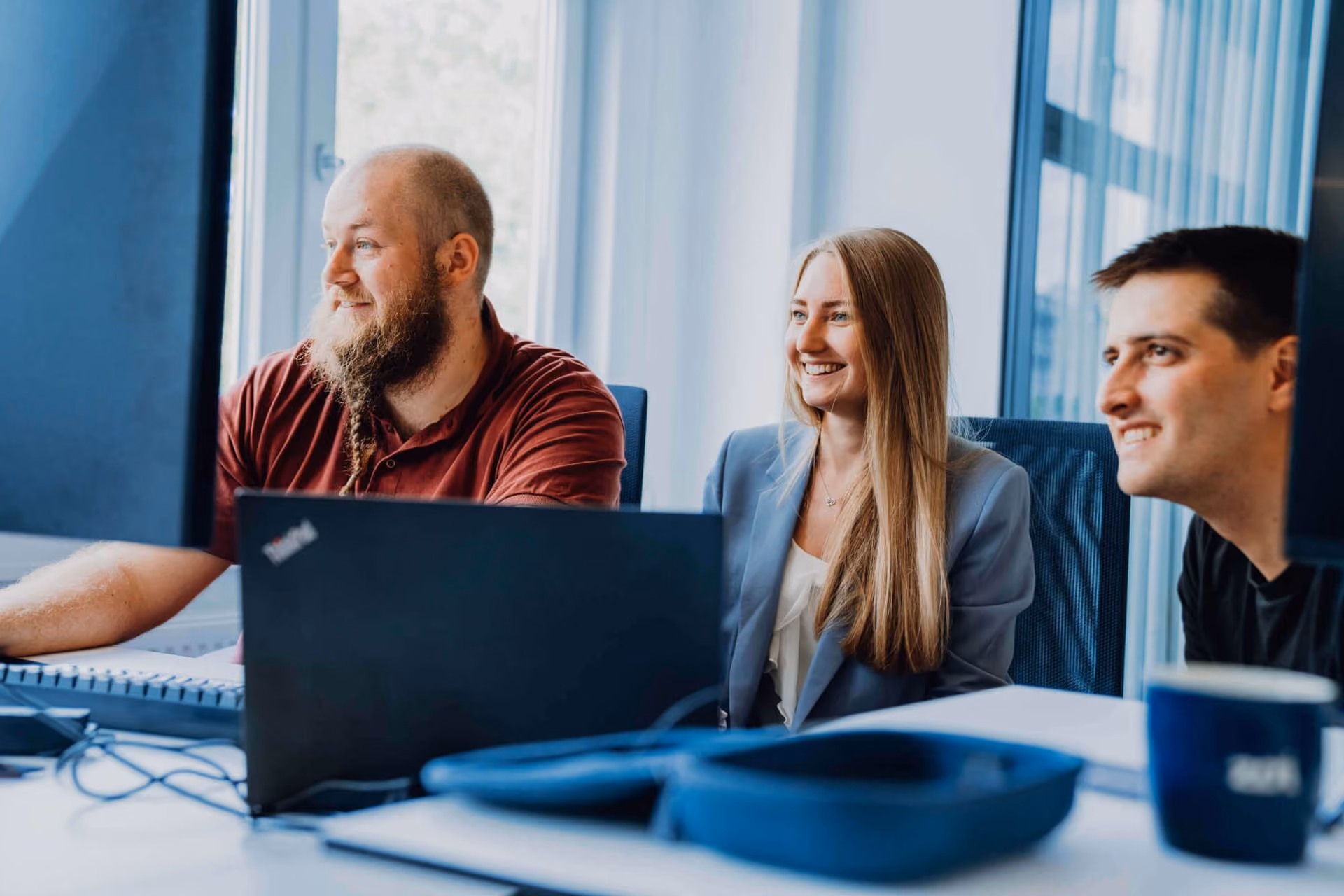 Three colleagues in an office smiling and looking at computer screens during a meeting.