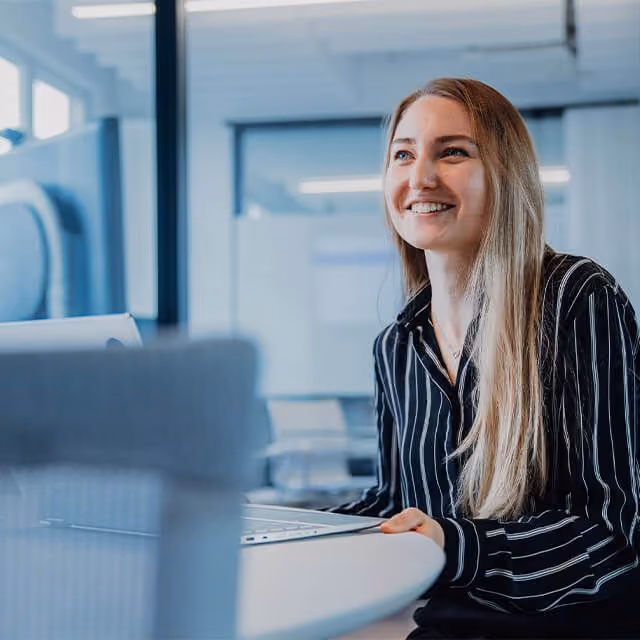 Smiling woman with long blonde hair and striped shirt sitting at a desk with a laptop in an office.