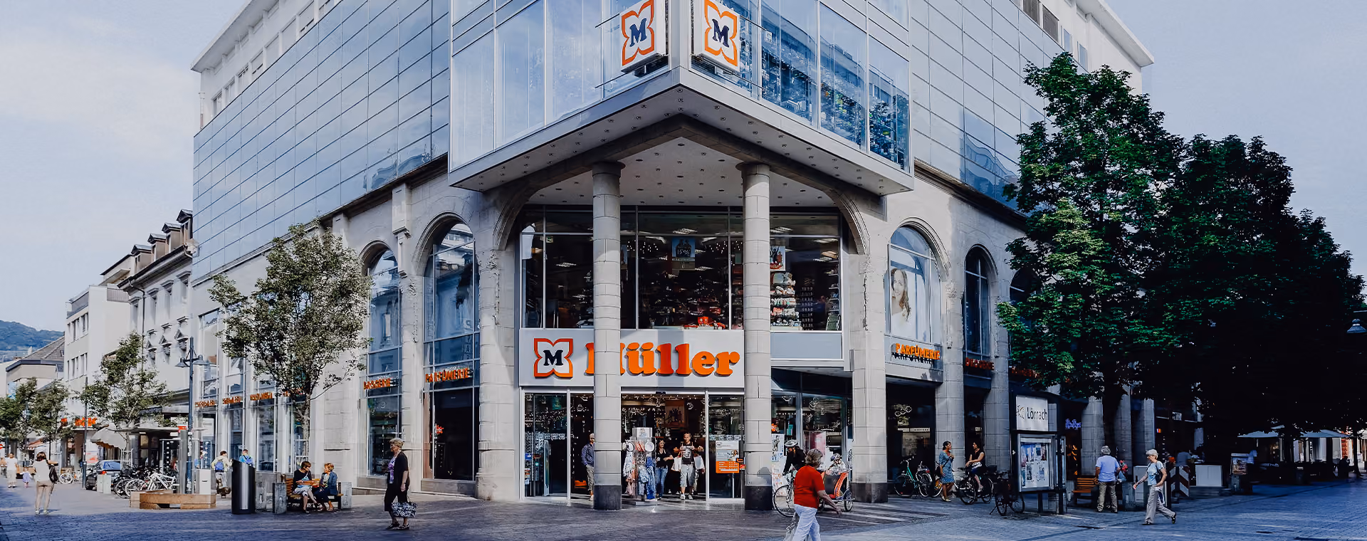 Modern corner store with large windows, 'Müller' signage, trees, and people walking nearby.
