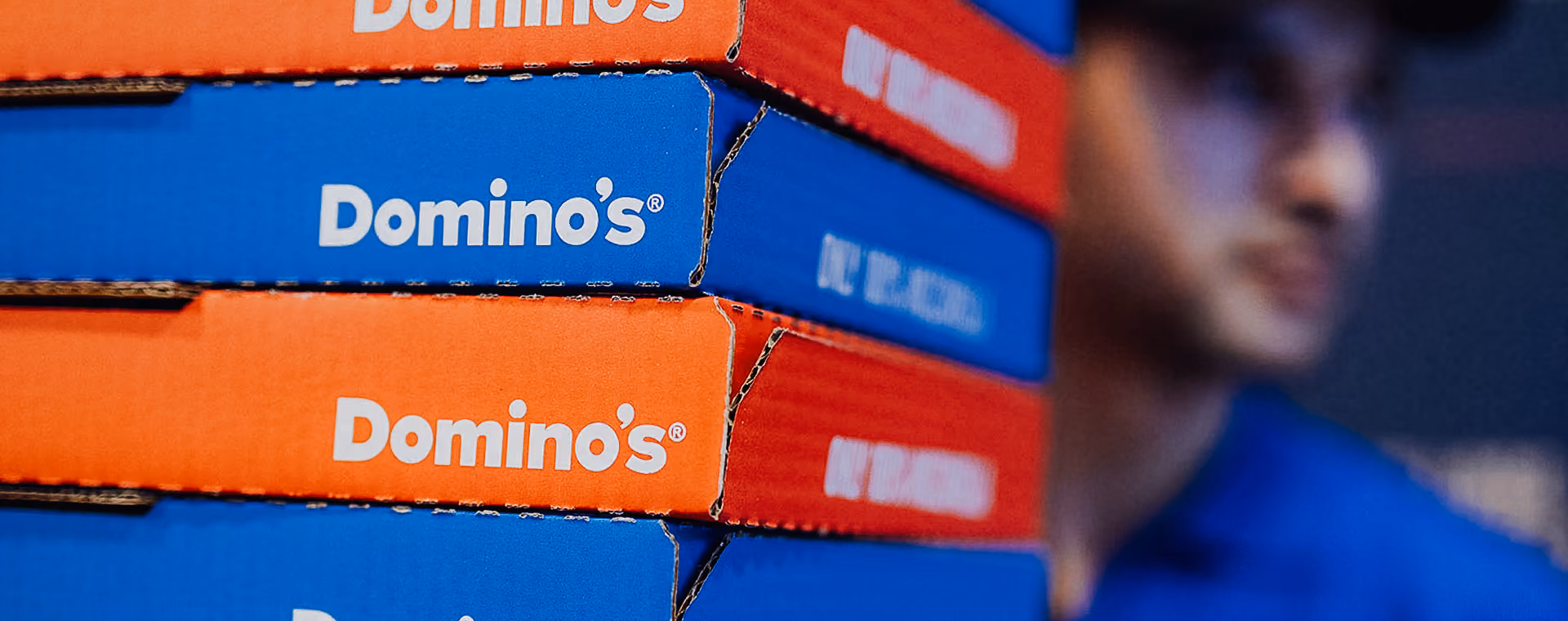 Stapel von Dominos Pizzaschachteln mit einer verschwommenen Person im Hintergrund.